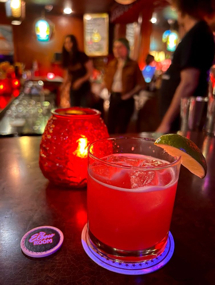 A pink cocktail in a tumbler glass served on the rocks with a fresh lime wedge. The cocktail is presented next to a glowing candle votive and poker chip with “The Elbow Room” logo. The items are positioned on a counter with three employees standing on the opposite side. 