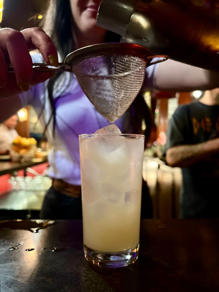 A bartender using a mesh strainer to complete the final touches on a pale yellow cocktail served on the rocks. 