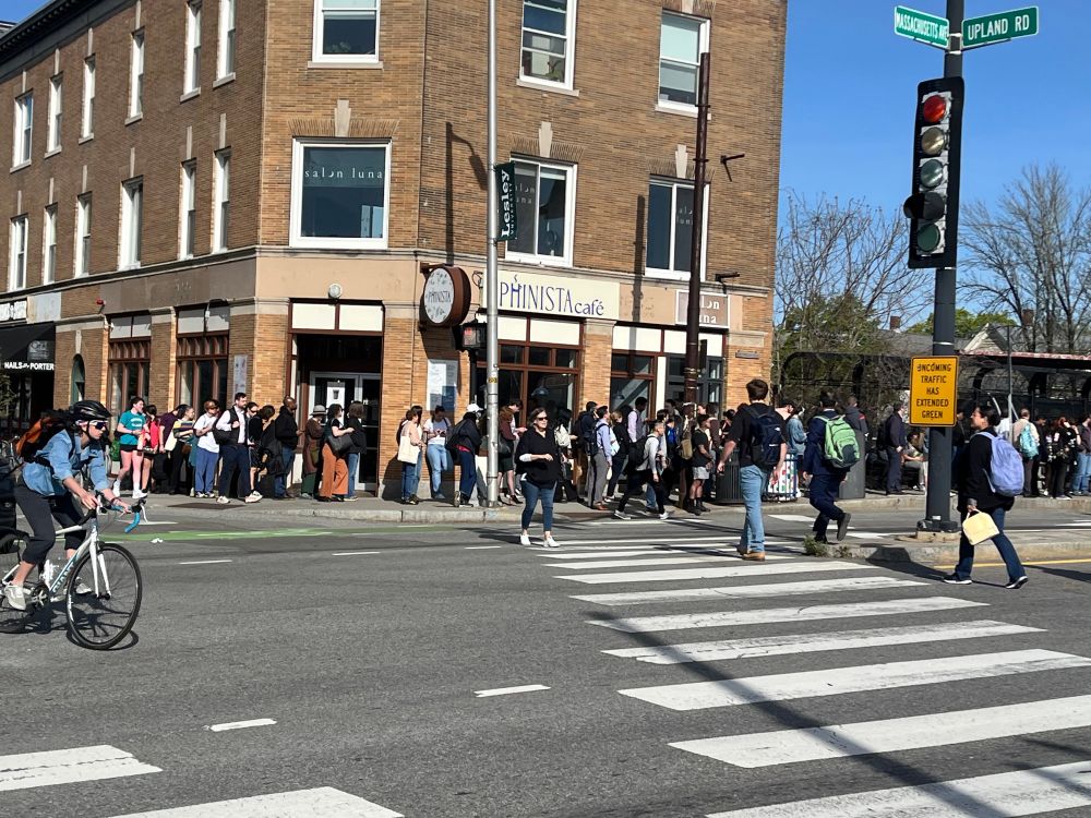 People waiting in line for a bus outside a 3 story brick building near a cross walk on Mass Ave in Cambridge ma