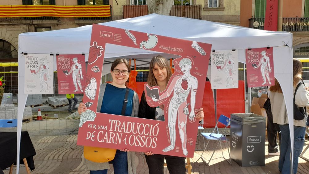 Dues dones en un photocall amb una figura humana anatòmica i el lema "Per una traducció de carn i ossos"
