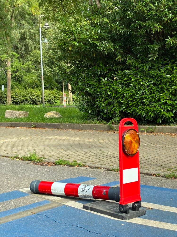 Umgefahrener Poller auf einer blau markierten Straße. Vor dem am Boden liegenden Poller ist ein Warnlicht postiert 