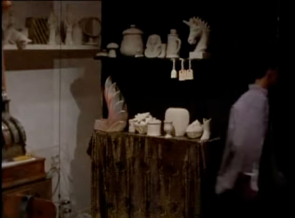 A screenshot from the mentioned episode: Ross, the main character of the episode, exits behind a curtain, showing a workshop with white figurines lining shelves in the background. There are jars, mugs, large chess pieces and an old timey cash register. 