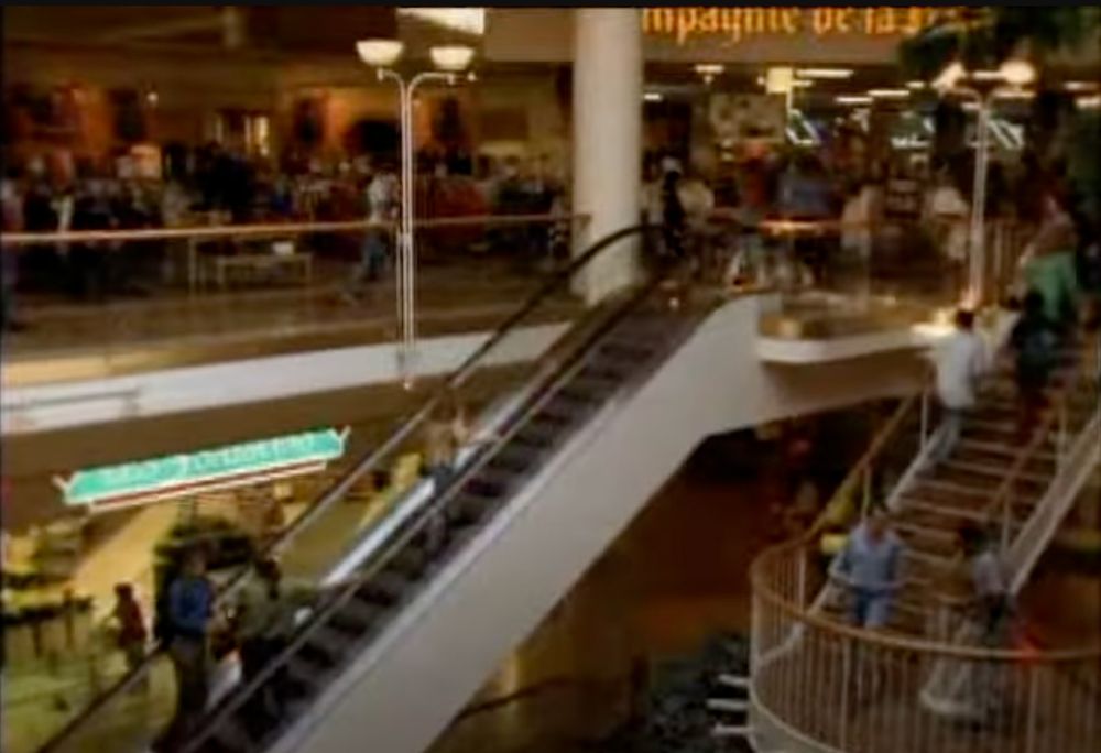 A screenshot of the establishing shot from the mentioned episode: a bustling multi-level mall, with a staircase and escalator featured prevalently in the middle. 