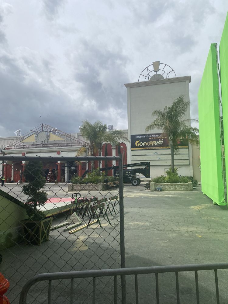 The parking lot of an abandoned movie theater, currently being reused to film a television show. A banner on the building says “Gomorrah”, and there is a large green screen to the right. 