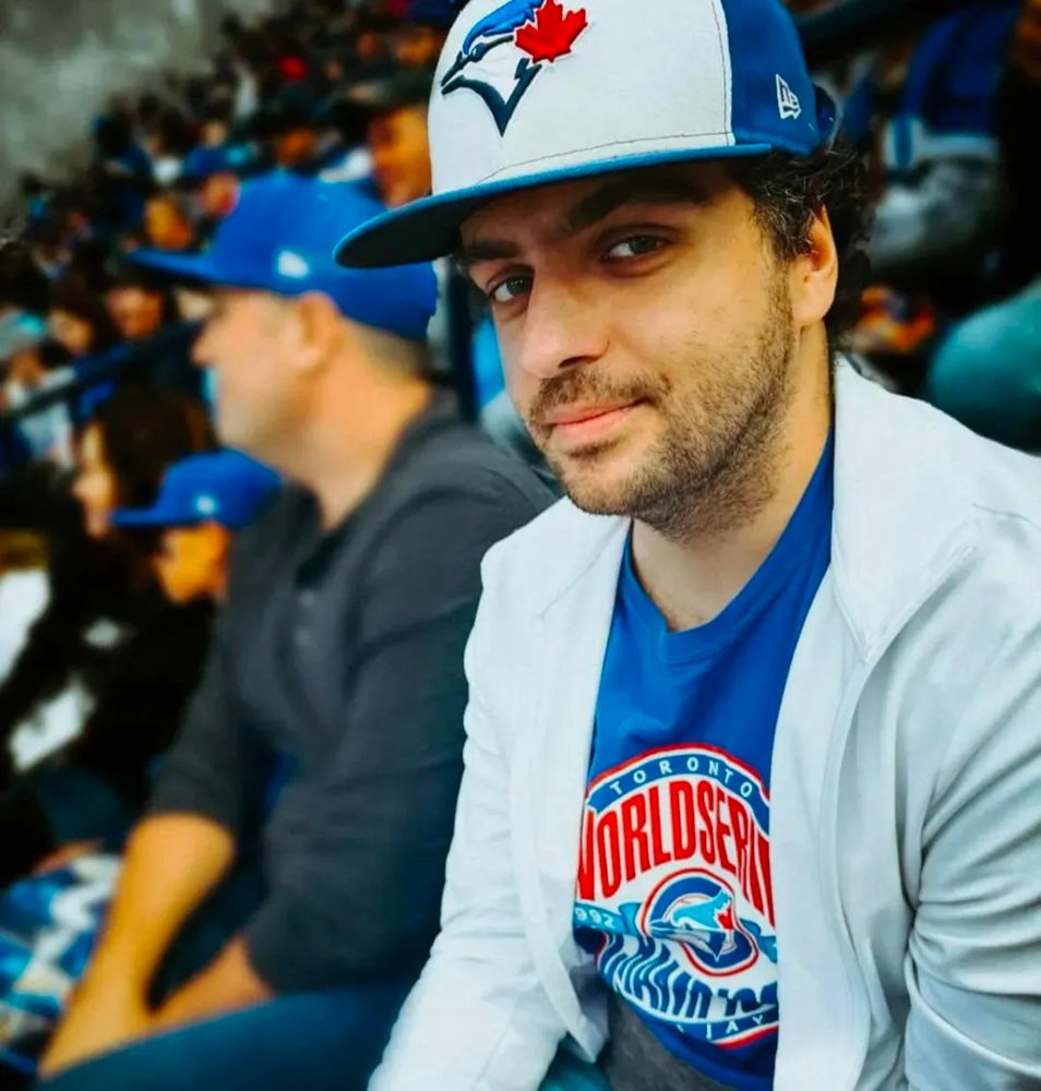Photo of me at a Jays game, half smiling at the camera while wearing a Jays hat, and a white zip up over a Jays tee