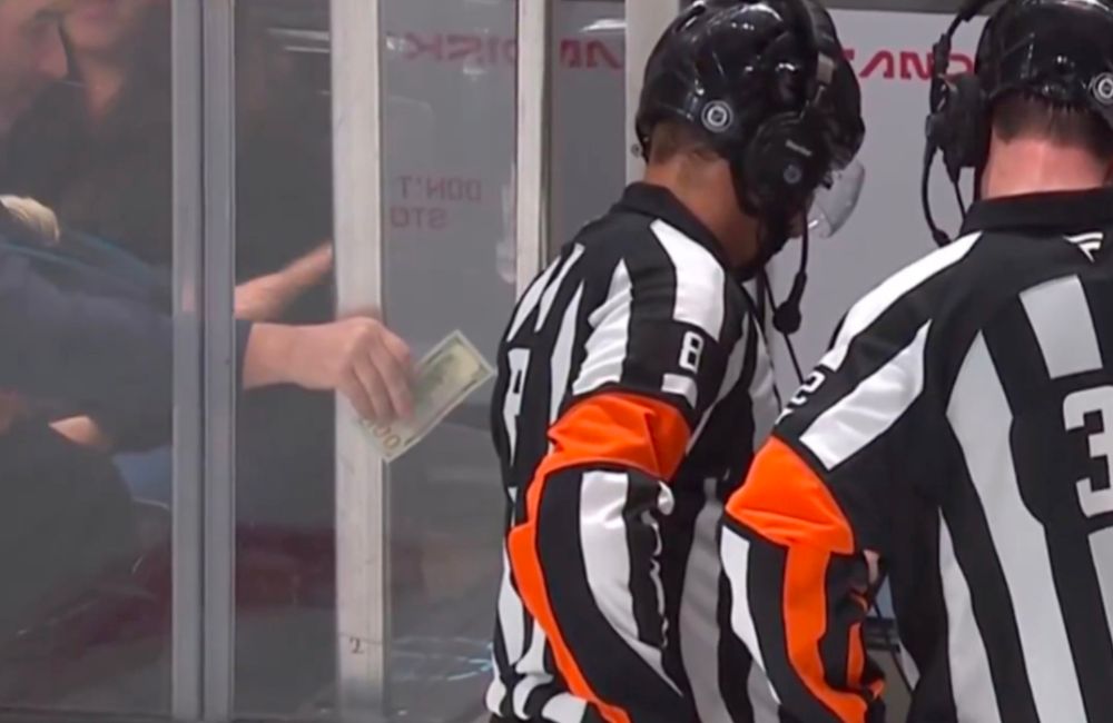 Two refs review a play on an off screen monitor as a fan reaches through the boards trying to hand the refs money. 