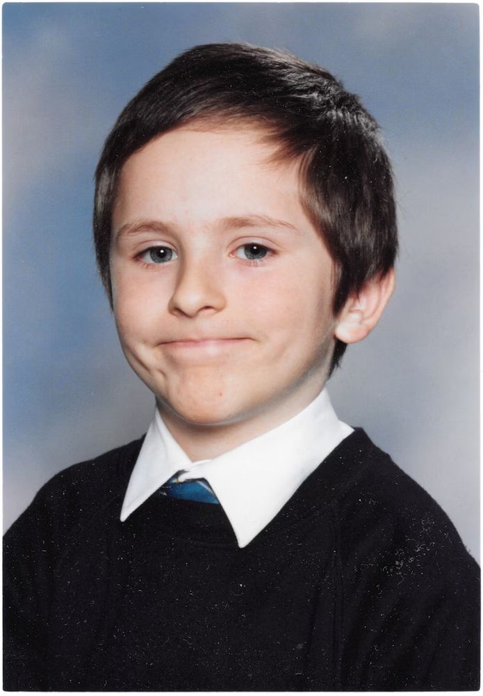A portrait of a young boy, wearing a tie and a sweater, making an awkward smile.