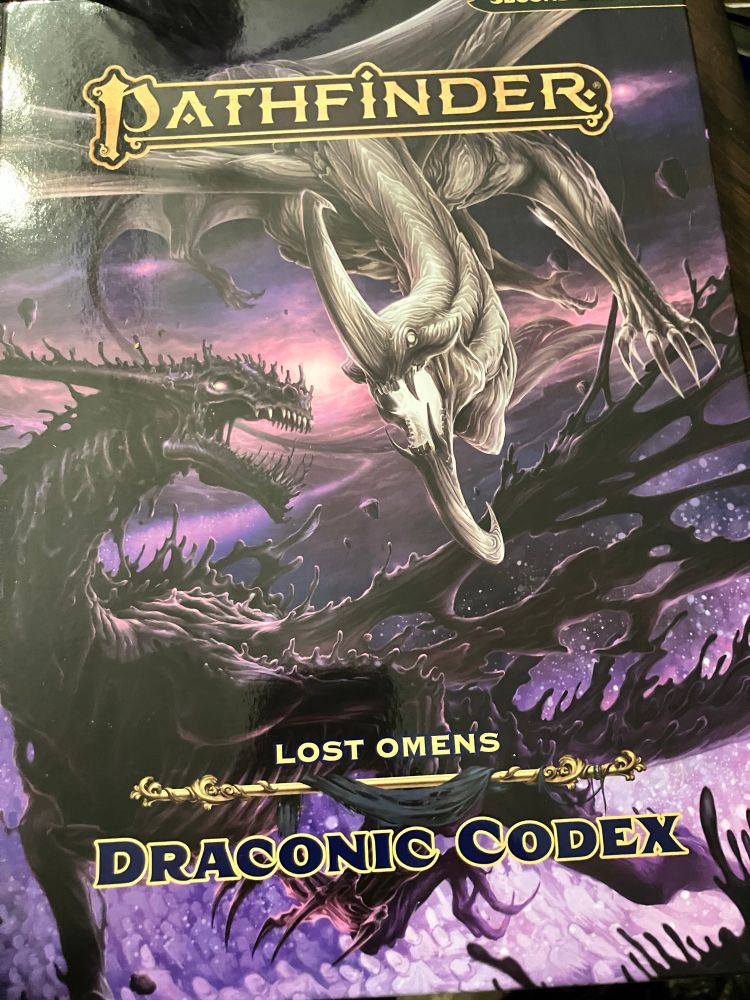 Gorgeous cover art of Pathfinder’s Draconic Codex, depicting two dragons: one a silver-grey Requiem Dragon, with matching horns curling back, one up from their forehead, the other down from their chin; the other a dark, partially-amorphous, skeletal-looking Resurrection Dragon. They circle each other before a deep purple “sky”, with a single brilliant, sun-like, light source in the distant background—the River of Souls, flowing from its origin point in Creation’s Forge. 
Art by Miguel Regodon Harkness