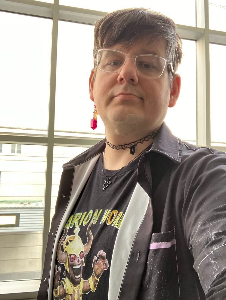A confident Raven, with hair styled to the right and looking reasonably nice, her usual trans necklace and d20 choker, and a dangly red stone on a gold earcuff. Same black and purple Fountain City Lodge button down over the Starfinder Golarion World t-shirt, with Tarby, the chibi Whispering Tyrant mascot. Her glasses are noticeably askew (stepped on; optom could get em back to wearable and functional, but no further—I kinda like the look, anyway)
