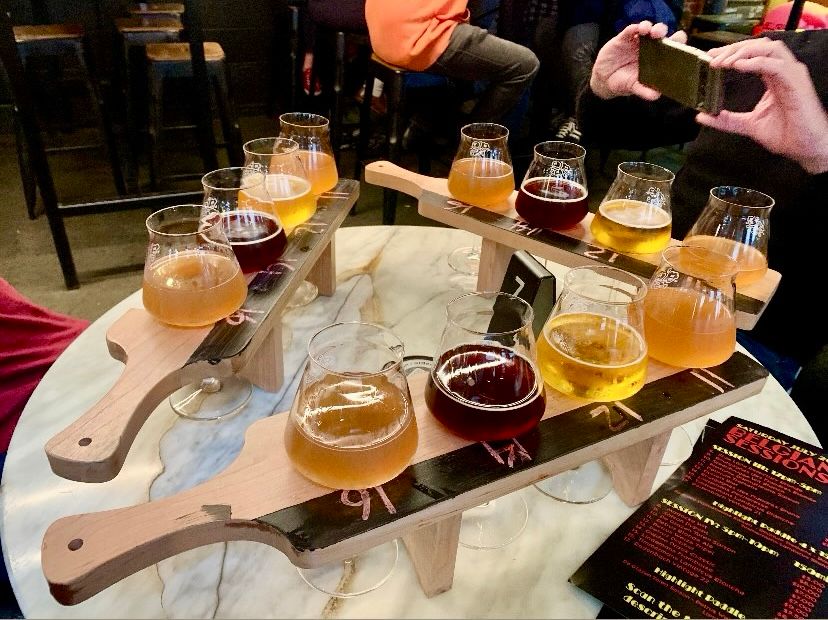scene from a beer tasting with glasses of different styles of beer nestled in paddles, there are three paddles each holding four glasses perched on a round table and formed into a kind of ‘tasting triangle’
