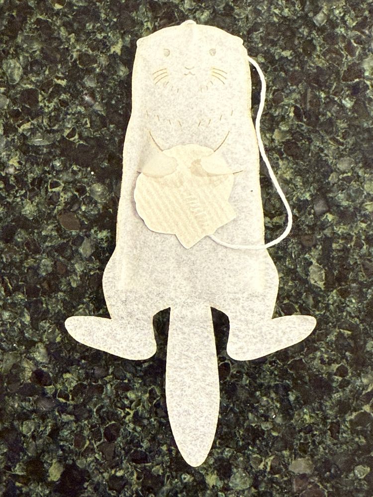 A tea bag shaped like a sea otter, complete with feet and a tail and a little face stamped on it. It has cutouts for its paws that allow it to hold the shell-shaped tag.
