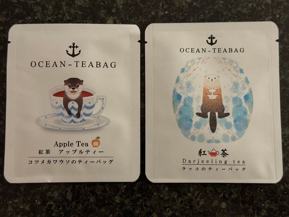 Two tea pouches containing apple flavored tea and Darjeeling, respectively. The apple tea has artwork of an Asian otter hanging out of a teacup while the Darjeeling has a sea otter laying on its back floating in the ocean holding on to a seashell.