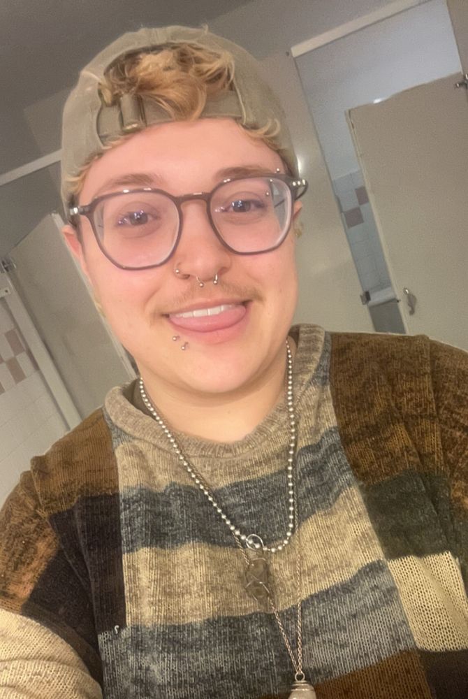 selfie of me, wearing a faded green backwards cap, color block earth tones sweater, and layered crystal necklaces. I have blond curls poking out from my hat, glasses, a septum and nostril piercings, two lip piercings, and an eyebrow ring visible
