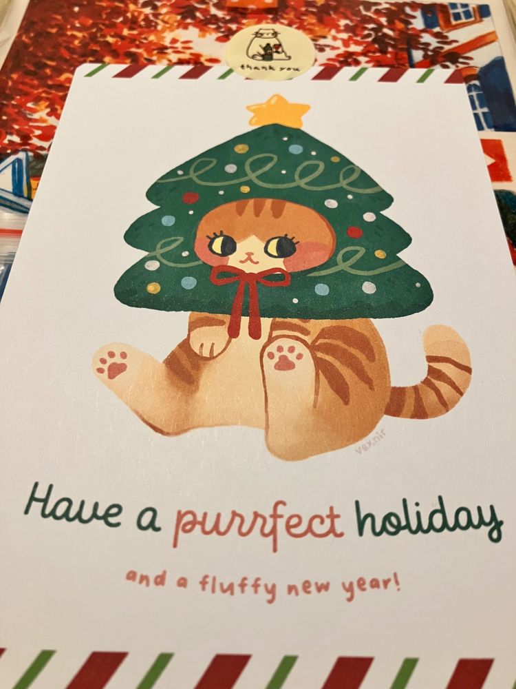 Holiday card saying ‘Have a purrfect christmas and a fluffy new year!’ Shows a cat with a christmas tree hat, from artist vexnir