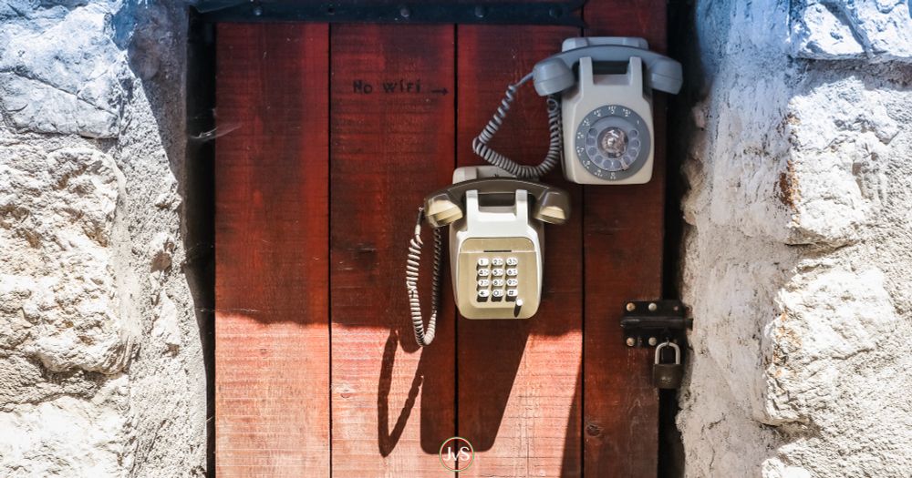 Eine rustikale Szene: In einer alten Steinmauer ist eine dunkelrote Holztür eingelassen. An der Tür sind zwei alte Festnetztelefone angebracht – eines mit Wählscheibe oben rechts, das andere mit Tastenfeld darunter. Über den Telefonen ist in schwarzer Schrift „No Wifi →“ zu lesen, was humorvoll auf die altmodische Kommunikation hinweist. Unten an der Tür hängt ein Vorhängeschloss. Das Bild vermittelt eine Mischung aus Nostalgie und augenzwinkerndem Kommentar zur modernen Technik.
