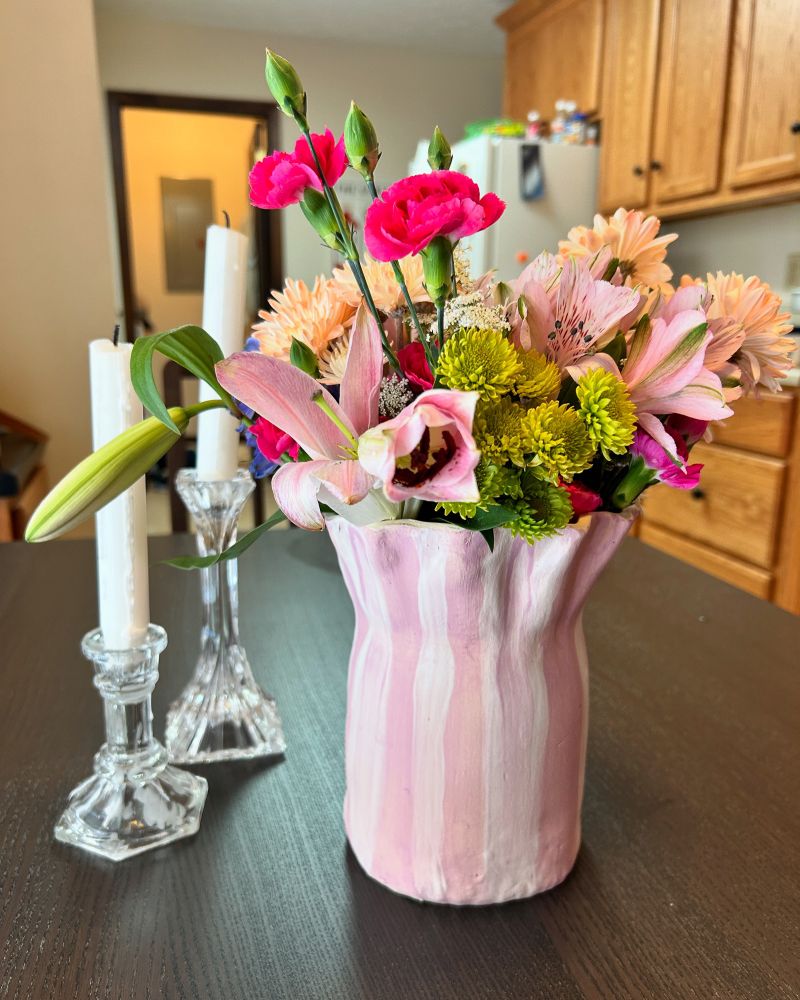 A purple and white striped vase full of flowers sits on a kitchen table beside 2 candles.