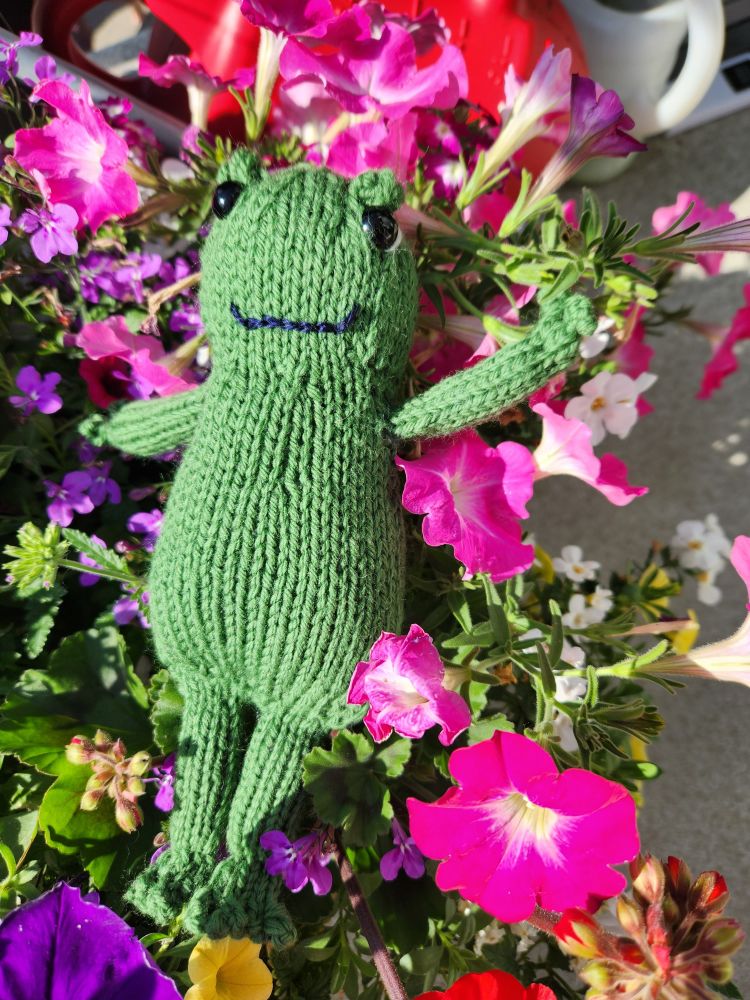 Knitted green frog relaxing in a pot of pink petunias.