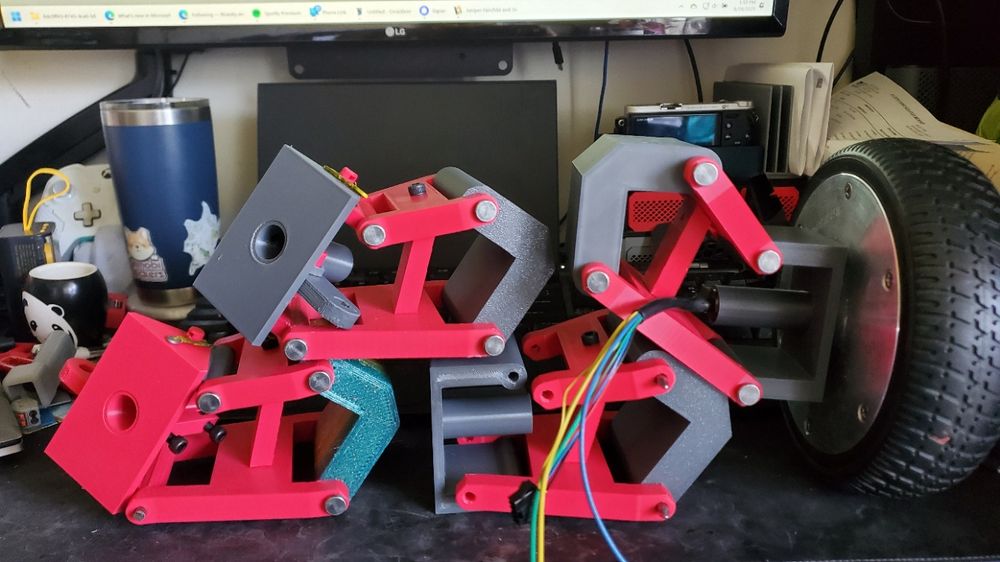 The image shows a partially assembled, brightly colored, 3D-printed robotic rover sitting on a dark surface next to a laptop and various other items.