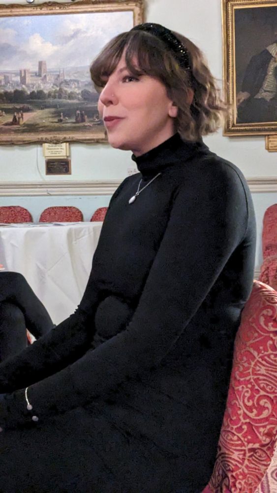 A white woman dressed in black with short wavy brown hair, sitting on a red brocaded chair.