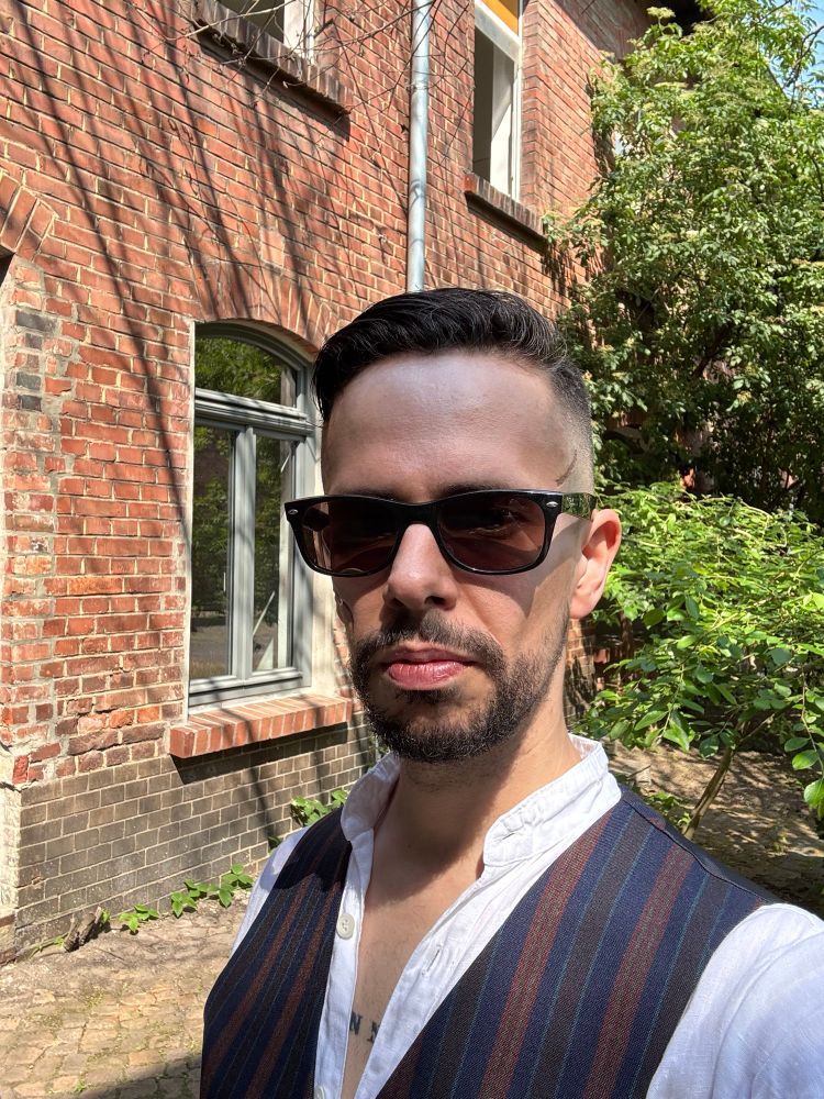 Man in a blue and brown vest wearing sunglasses and a white linen shirt standing in front of a brick building.