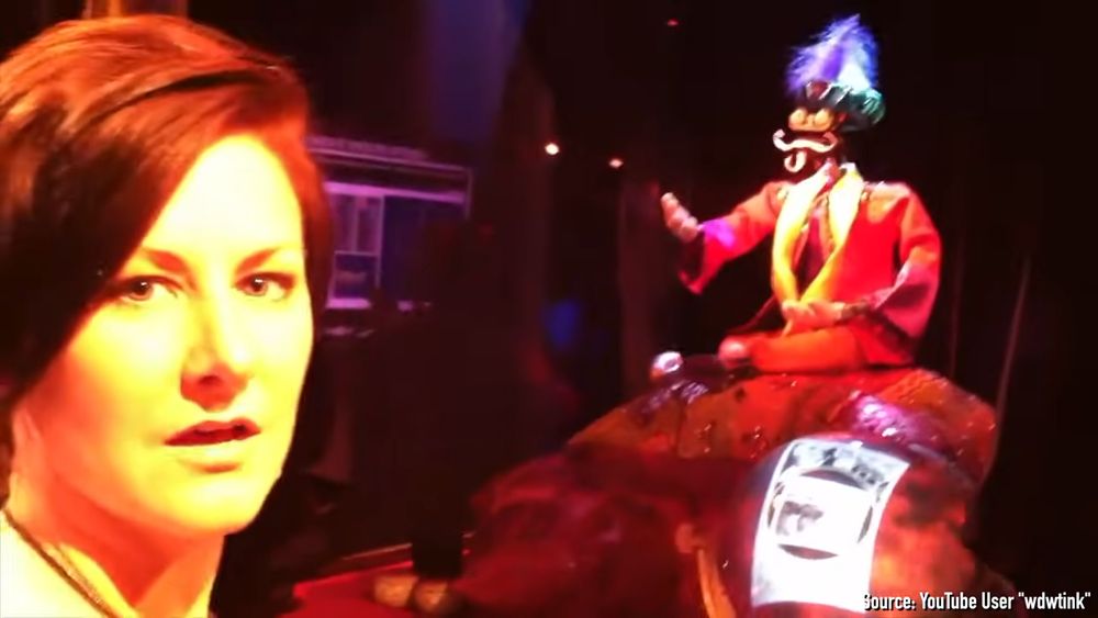 a woman looks into the camera with an exasperated and unimpressed expression while standing before a small animatronic fortune teller