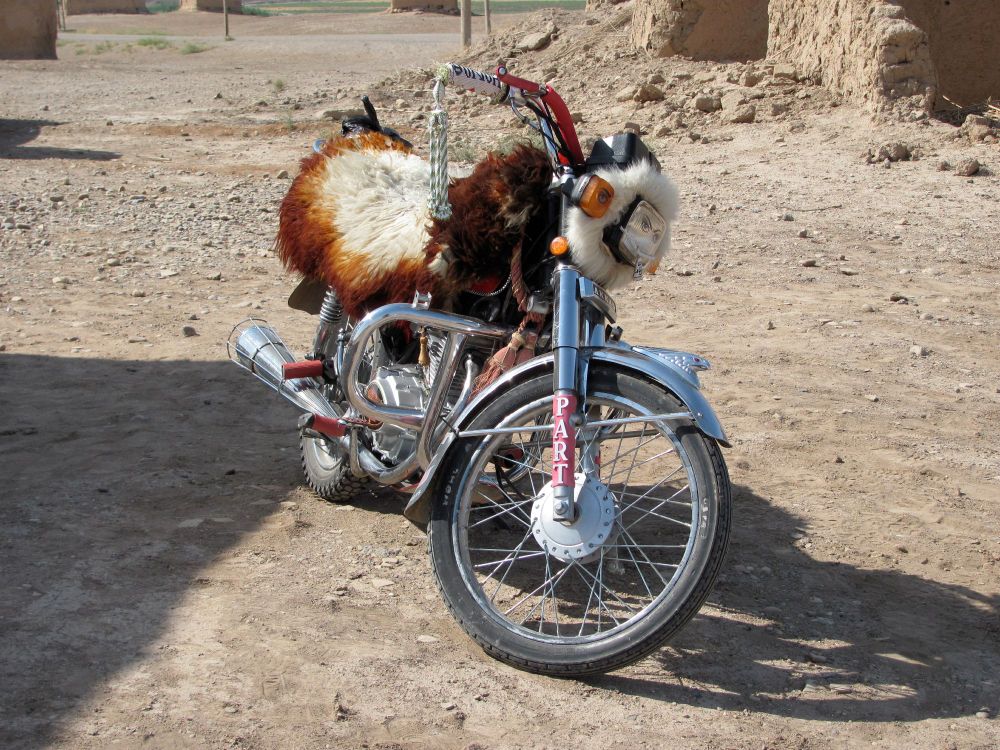 Motorcycle covered in goat fur with a red strut that has the word "PART" on it in capital letters. The bike is parked on desert ground in the middle of a Syrian archaeological excavation site (photo taken in summer of 2010).