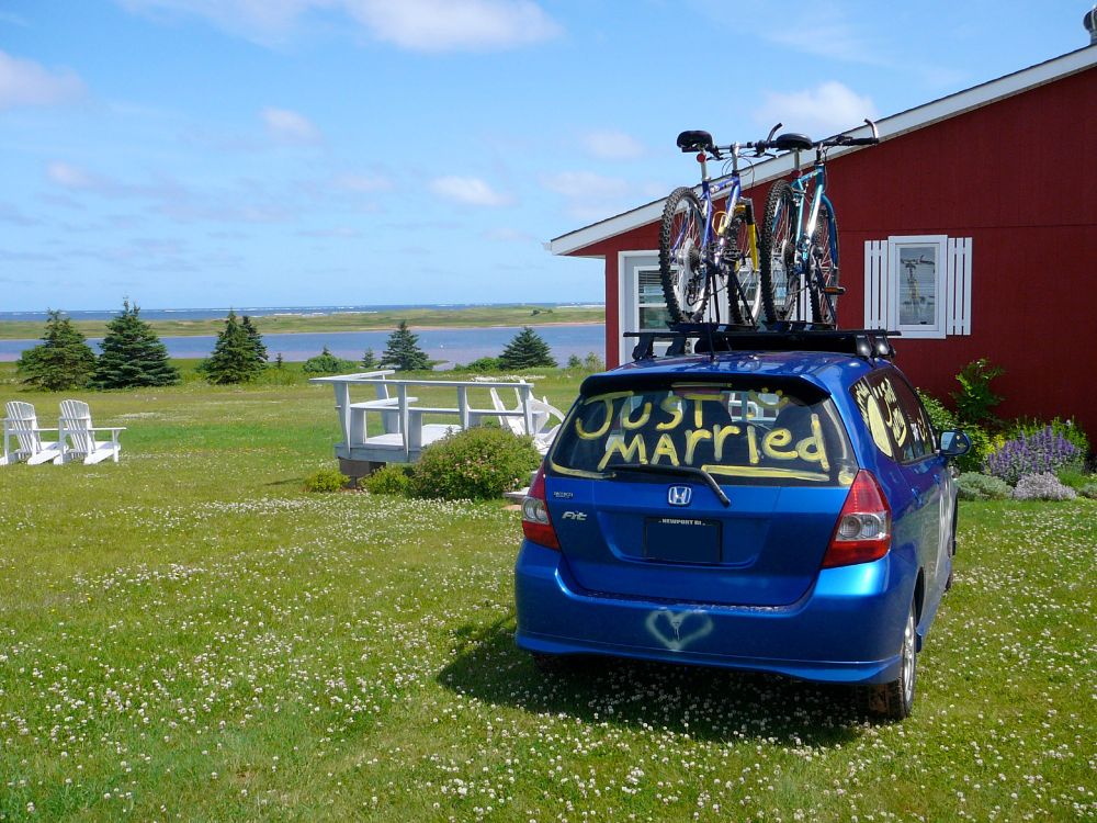 Blue car with bikes on the roof and "Just Married" written on the back window parked in a grassy field in front of a red cottage, ocean/bay in the distance. 