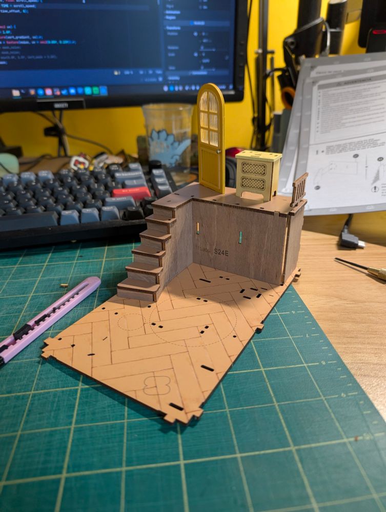 Wood/cardboard model progress (base, small staircase going up to arched yellow door) on a cutting mat with computer keyboard and instructions in the background. 