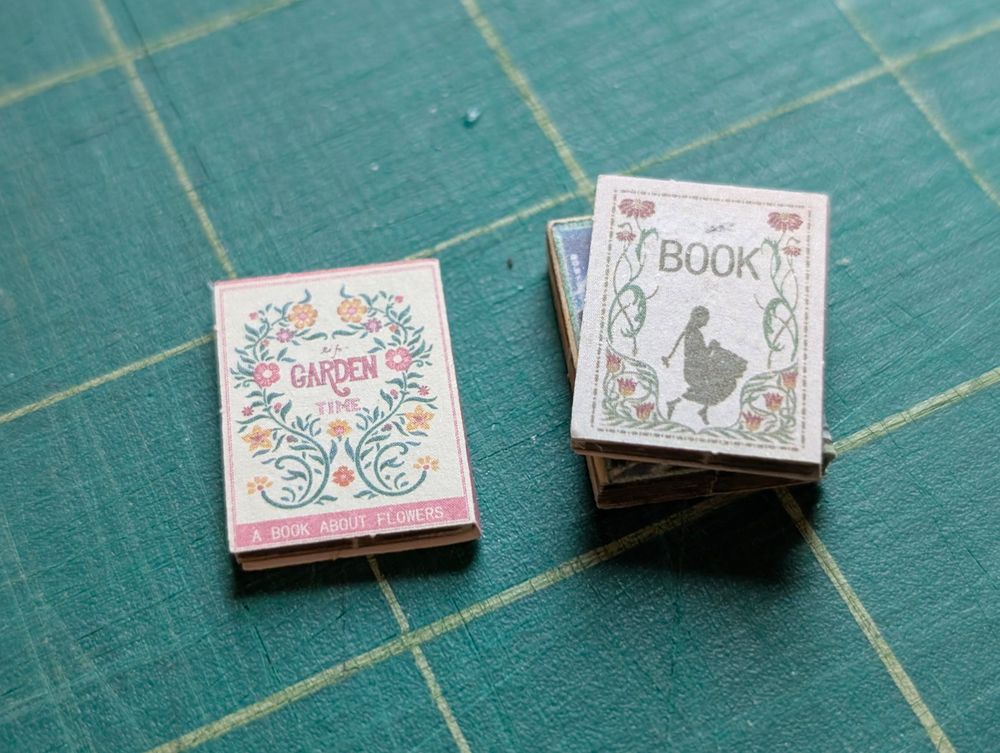 two small fake books made of paper and balsa wood on a cutting mat. one is titled "Garden Time, a Book About Flowers" and the other just "Book" with a girl silhouette on it. 