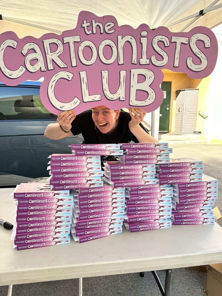 Iz - a white person with short dirty-blond hair wearing a black t-shirt - behind stacks of The Cartoonists Club on a table holding up a large purple sign that matches the title font and reads “The Cartoonists Club”. 