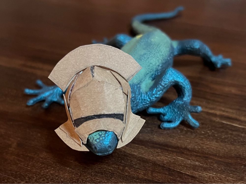 Small greenish blue tit lizard on a dark wood table, wearing a cardboard helmet modeled after the orc helmets in Lord of the Rings.