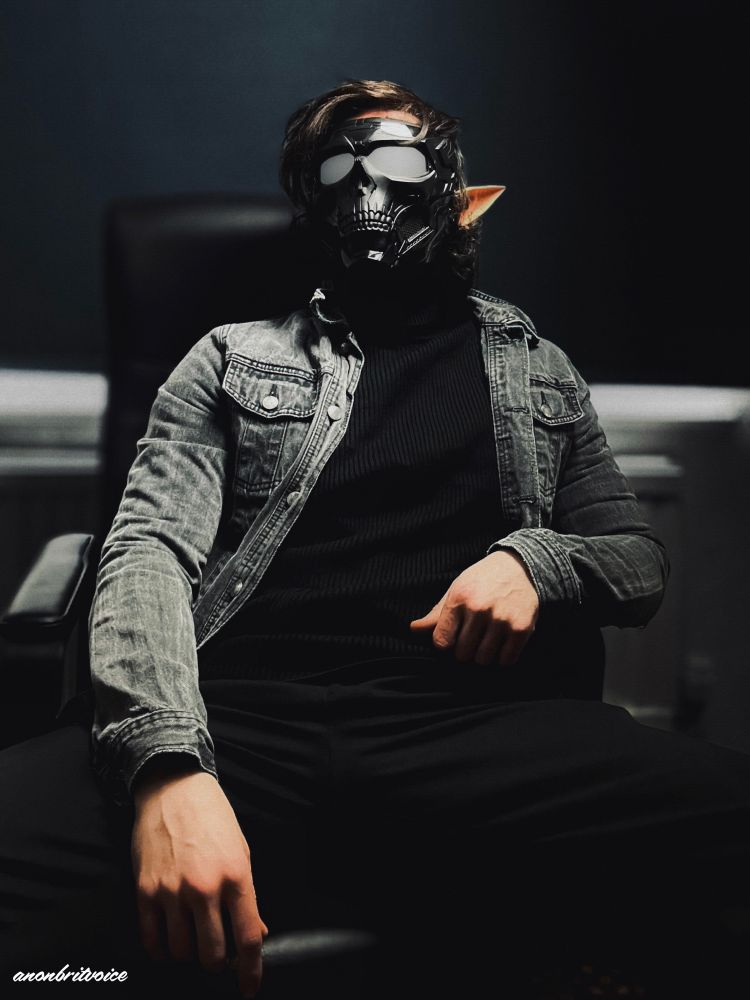 Photo of a man wearing a black trousers, black jumper, grey denim jacket, skull mask and fae ears, siting in a chair with legs wide