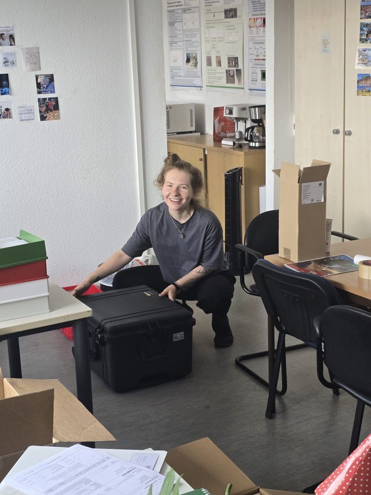Office room, a young woman holding a black case in their hands, smiling at the camera | Büroraum, eine junge Frau hält einen schwarzen Koffer in der Hand und lächelt in die Kamera
