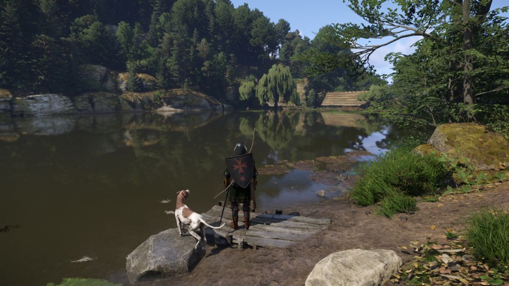 From Kingdom Come: Deliverance II - Henry with teh dog Mutt standing on the edge of a small lake and looking towards the forest.