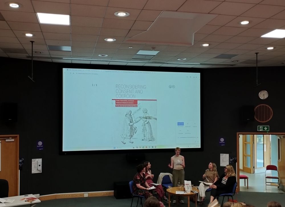In a large lecture theatre, five women are gathered around a small coffee table engaged in discussion in front of an audience. Behind them is a large screen displaying the cover of a book. The book's title is: 'Reconsidering Consent and Coercion - Power, Vulnerability, and Sexual Violence in Medieval Literature'. 