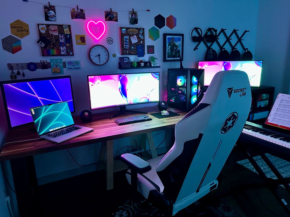 A wooden desk in a dimly lit room. A couple monitors, gaming chair, keyboard piano, various decorations on the wall, RGB lighting.