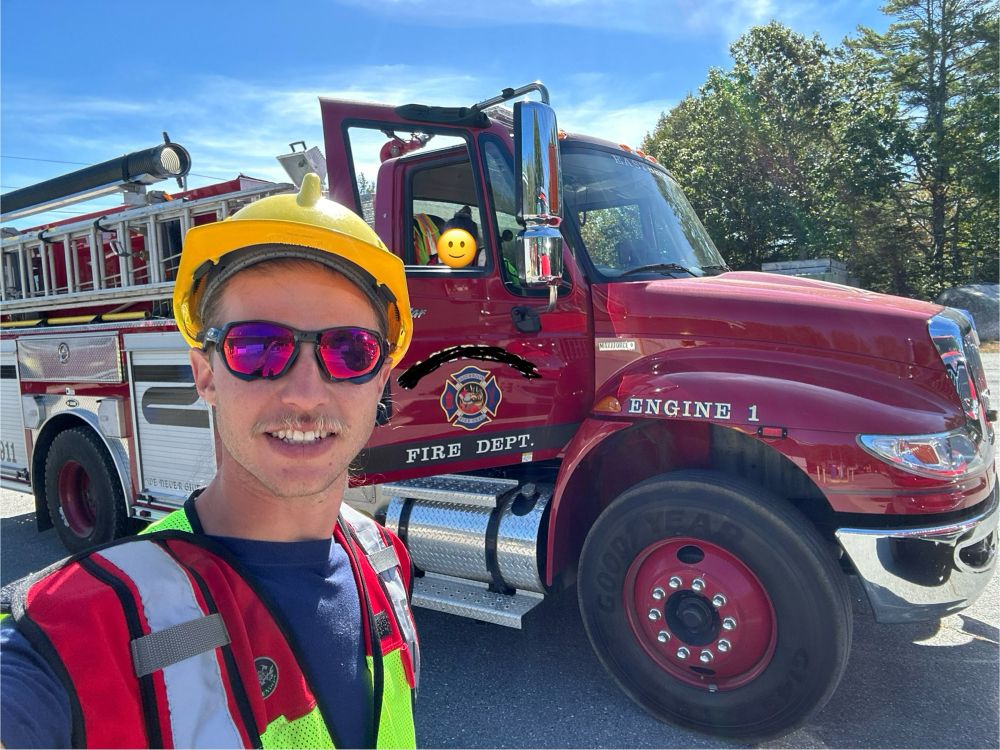 Jake is standing in front of a fire apparatus labeled “Engine 1”. They have a red and neon green reflective vest on with a yellow helmet and red sunglasses.