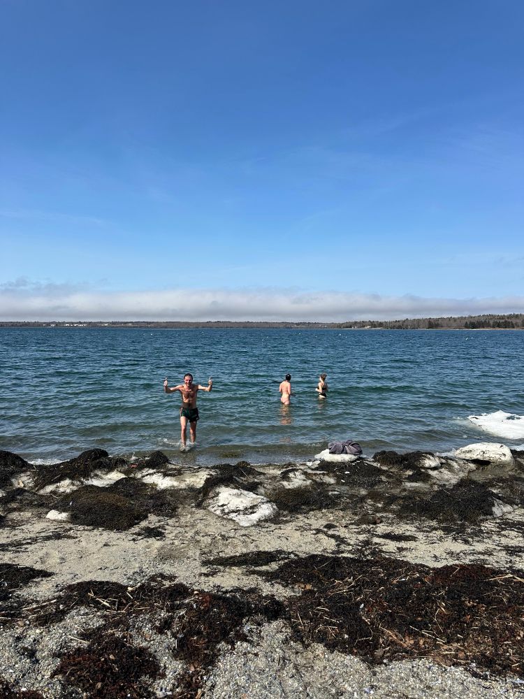 Jake is giving two thumbs up as they exit the ocean.  The foreground is an ice and seaweed covered beach. A mostly clear blue sky is overhead. 