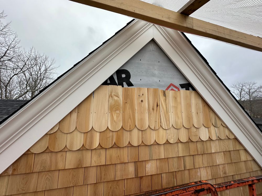 Two rows of shingles with rounded bottoms are nailed above courses of square shingles. There is white trim meeting at the peak of a gable on a house.