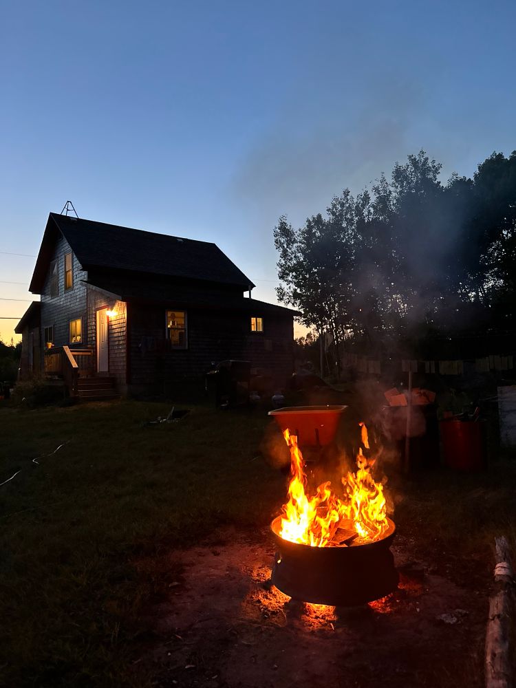 A cute cottage is silhouetted against the afterglow of sunset, surrounded by shadows of trees. A small fire blazes in the foreground, casting a warm orange glow on the ground.