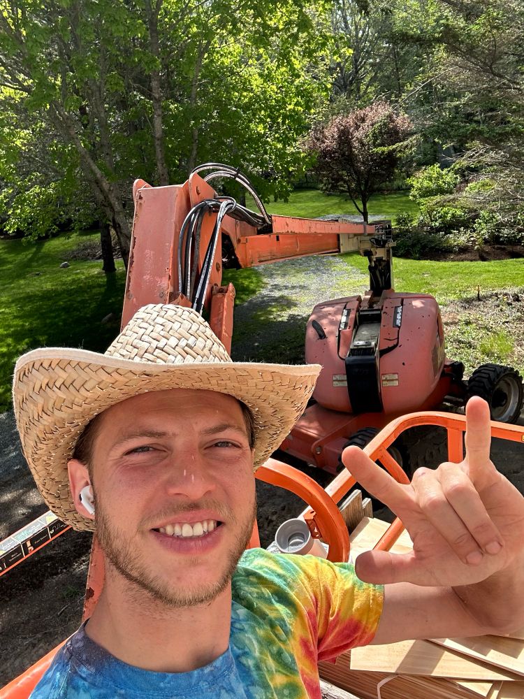 Jake is in the basket of a boom lift making the sign for “I love you”. They are wearing a tie dye shirt and straw western hat and have an AirPod in their ear.
