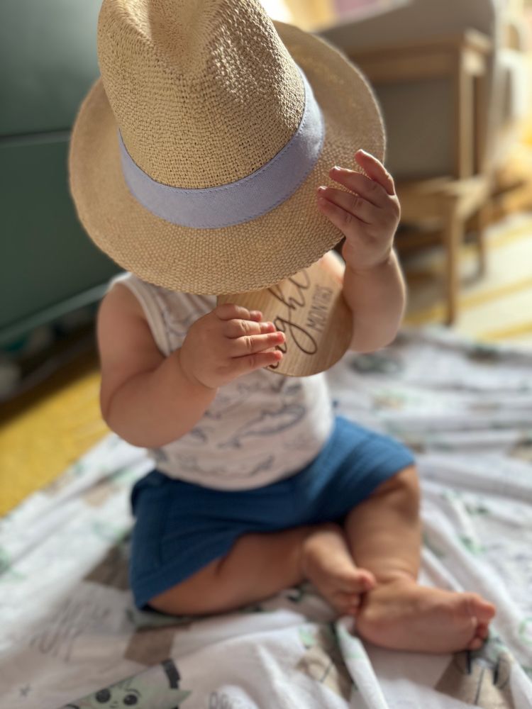 Photo shows a baby with his face covered by a straw hat holding a wooden medallion that says “eight months”