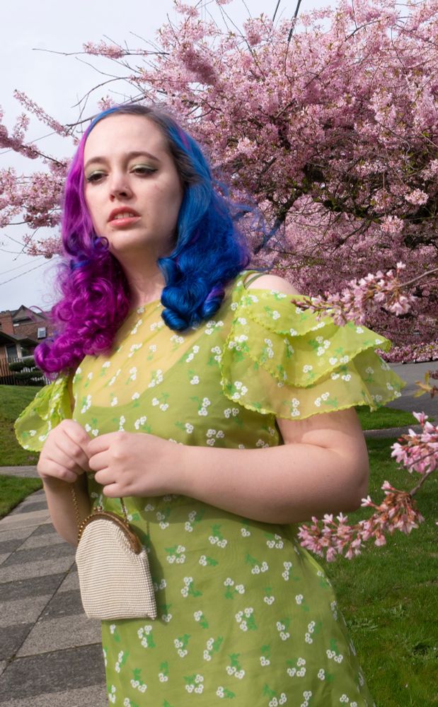 Haley a white femme with half blue half pink hair wearing showing a closeup of a the fluttery scalloped sleeves of 1930s dress. She is holding a small white beaded purse. There are pink cherry blossom trees and grass in the background
