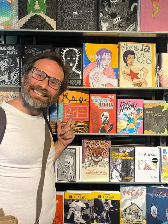 A very happy Henry Widdicombe pointing at his comic, How to Survive an Affair on the shelves of Gosh Comics in amongst loads of very cool looking comics 