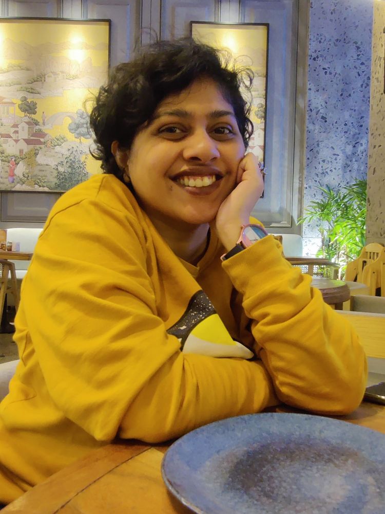 Astralsoul wearing a ochre yellow sweatshirt, leaning forward against a dining table, supporting their cheek by their left fist. a gray plate is in front of them on the table and the wall behind is grey with large gold accent mural paintings. their smile is wide and they look happy.
