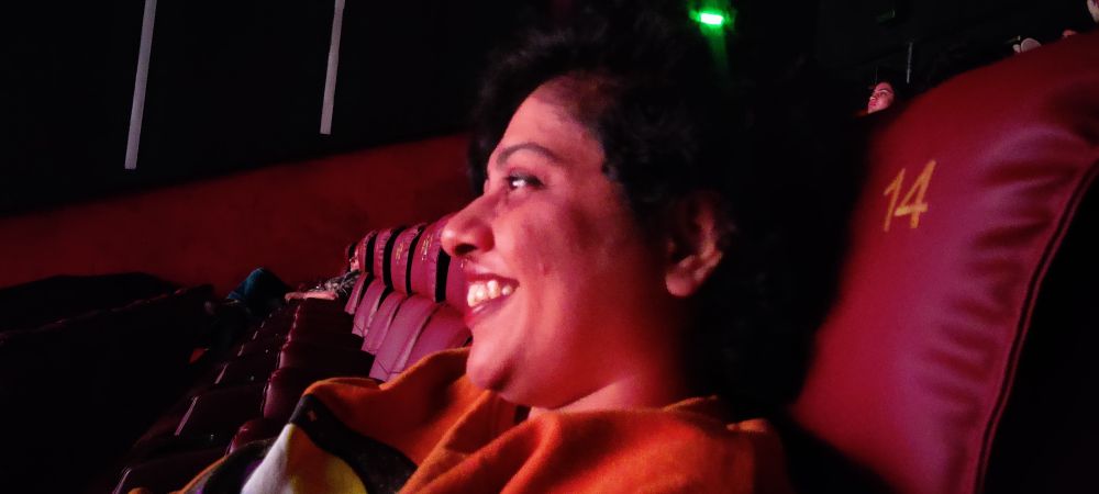 Astralsoul is seated in a cinema, their maroon seat is marked with the number 14. next to them is a row of empty seats. their face is lit by the screen in front of them and glee on their face is a function of watching Kal Ho naa ho in theatres after all these years. 