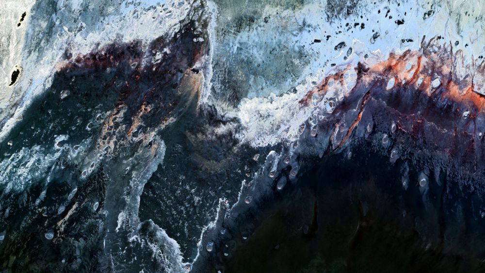A satellite picture of a detail from the shore of a lake showing a dramatic and colorful landscape with swirling patterns of dark blue, white, and rusty red.