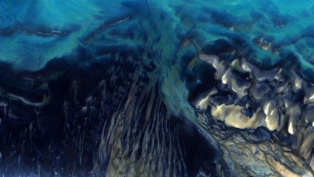 A satellite picture of the seawater on a coastline, with dark blue and teal tones interweaving in abstract patterns.