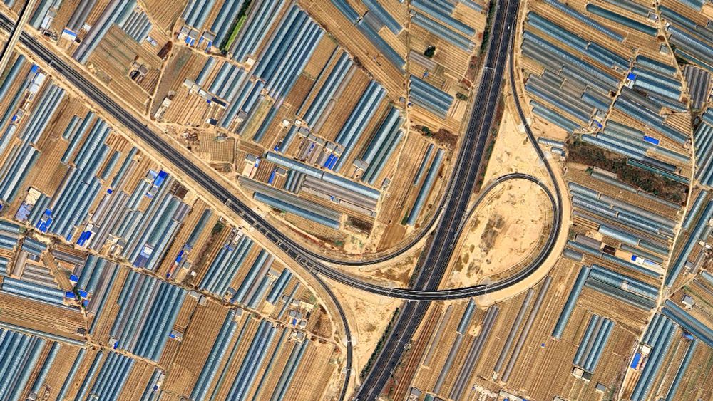 a satellite picture of an intensive agricultural landscape with series of grey-blueish greenhouses on light brown soil. the landscape is traversed by a highway
