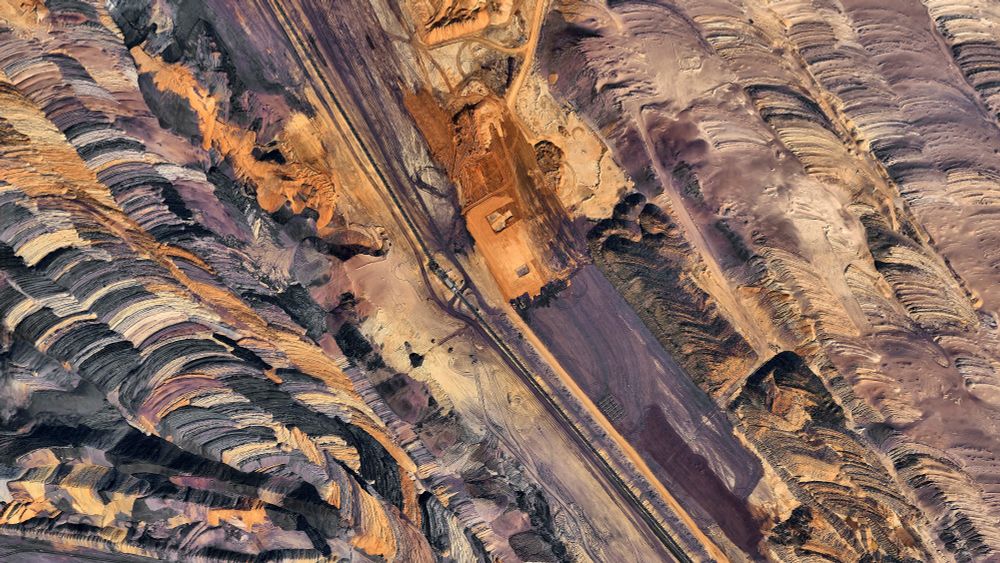 A satellite picture of a large open-pit mine with terraced layers of excavated earth in colors like purple, black, orange, and yellow. A flat central area contains buildings and equipment, connected by roads across the site. The stepped terrain reveals geological strata shaped by industrial mining.